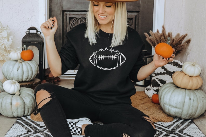 Puede incluir: Sudadera negra con el texto "gameday" sobre un gr&aacute;fico de f&uacute;tbol americano blanco. La imagen presenta calabazas y decoraci&oacute;n oto&ntilde;al, lo que sugiere un producto tem&aacute;tico de temporada o deportivo.