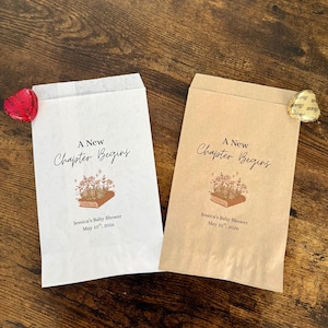 May include: Two paper favor bags, one white and one tan, with the text "A New Chapter Begins". Each bag has a floral book illustration and the text "Jessica's Baby Shower May 10th, 2026". A heart-shaped candy and a gold foil candy are visible.