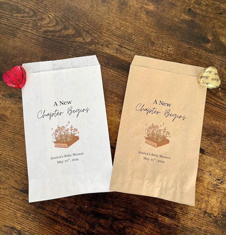 May include: Two paper favor bags, one white and one tan, with the text "A New Chapter Begins". Each bag has a floral book illustration and the text "Jessica's Baby Shower May 10th, 2026". A heart-shaped candy and a gold foil candy are visible.