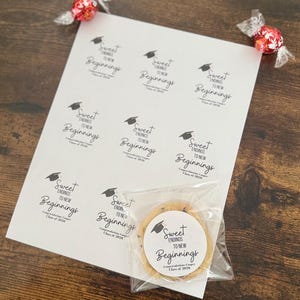 K&ouml;nnte beinhalten: Ein Bogen wei&szlig;er Aufkleber mit dem Text "Sweet Endings to New Beginnings" und einer Grafik einer Graduiertenkappe. Ein Schokoladen-Chip-Keks in einer durchsichtigen T&uuml;te befindet sich neben den Aufklebern. Zwei rote Bonbons in Klarsichtverpackungen sind ebenfalls im Bild.