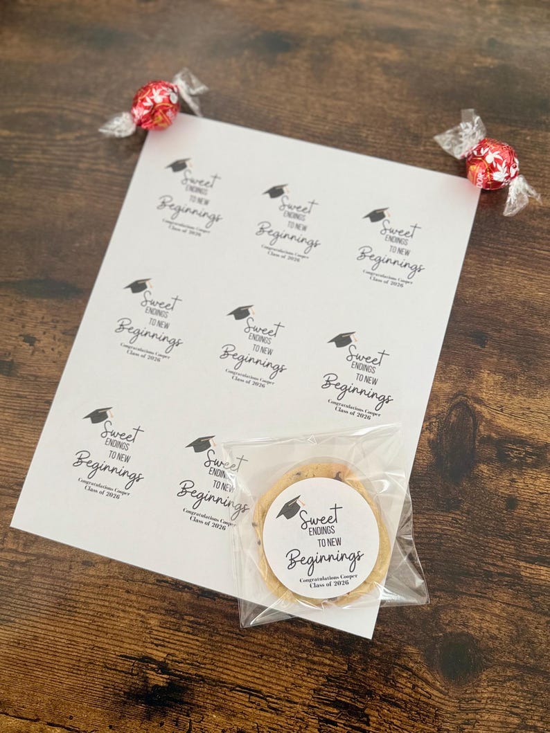 K&ouml;nnte beinhalten: Ein Bogen wei&szlig;er Aufkleber mit dem Text "Sweet Endings to New Beginnings" und einer Grafik einer Graduiertenkappe. Ein Schokoladen-Chip-Keks in einer durchsichtigen T&uuml;te befindet sich neben den Aufklebern. Zwei rote Bonbons in Klarsichtverpackungen sind ebenfalls im Bild.