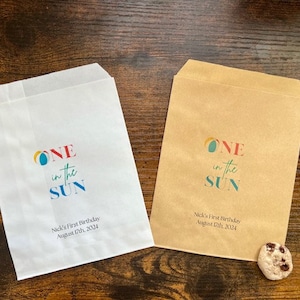 May include: Two paper treat bags, one white and one brown, with the text "ONE in the SUN" in colorful letters. The bags are for Nick's First Birthday, August 17th, 2024. A chocolate chip cookie rests near each bag.