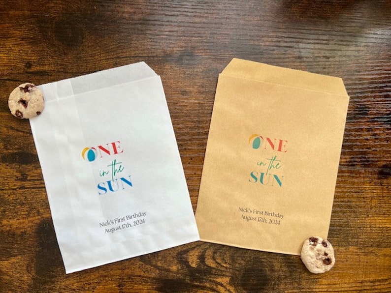 May include: Two paper treat bags, one white and one brown, with the text "ONE in the SUN" in colorful letters. The bags are for Nick's First Birthday, August 17th, 2024. A chocolate chip cookie rests near each bag.