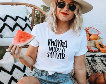 May include: White t-shirt with the text "MAMA NEEDS A SELTZER" in black script. The person is holding a watermelon slice. The background includes a wicker chair, decorative pillows, and a bowl of fruit.
