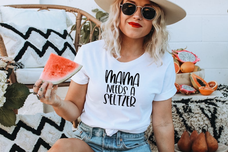 Puede incluir: Camiseta blanca con el texto "MAMA NEEDS A SELTZER" en escritura negra. La persona sostiene una rodaja de sand&iacute;a. El fondo incluye una silla de mimbre, cojines decorativos y un cuenco de fruta.