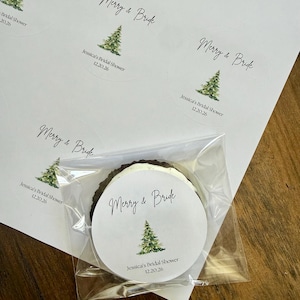 May include: A sheet of circular white stickers featuring a watercolor green Christmas tree illustration, the text Merry & Bride, and the date 12.20.26. One sticker is applied to a round dark cookie wrapped in a clear plastic cellophane bag.