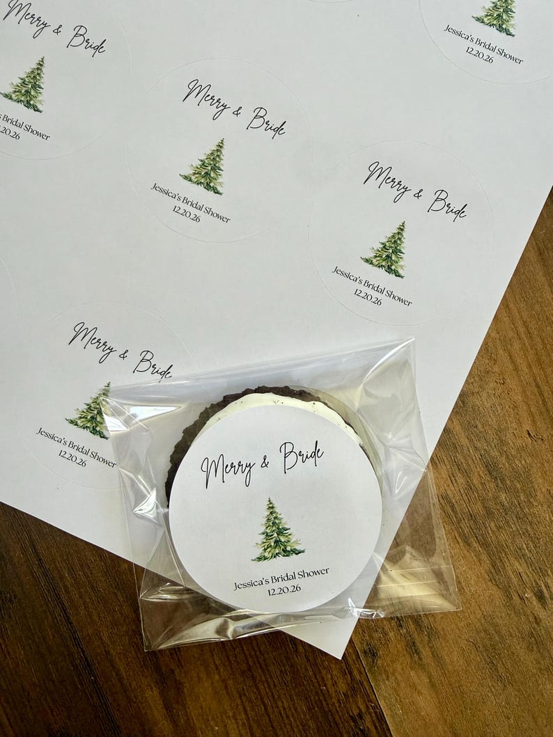 May include: A sheet of circular white stickers featuring a watercolor green Christmas tree illustration, the text Merry & Bride, and the date 12.20.26. One sticker is applied to a round dark cookie wrapped in a clear plastic cellophane bag.