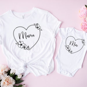 May include: White t-shirt and onesie set. The t-shirt features the word "Mama" inside a heart-shaped floral design. The onesie has the word "Mini" in a similar heart-shaped floral design. The set is displayed on a pink background.