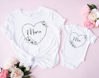 May include: White t-shirt and onesie set. The t-shirt features the word "Mama" inside a heart-shaped floral design. The onesie has the word "Mini" in a similar heart-shaped floral design. The set is displayed on a pink background.