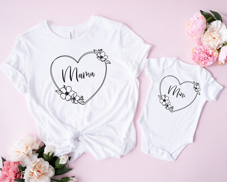 K&ouml;nnte beinhalten: Wei&szlig;es T-Shirt und Body-Set. Das T-Shirt zeigt das Wort "Mama" in einem herzf&ouml;rmigen Blumendesign. Der Body hat das Wort "Mini" in einem &auml;hnlichen herzf&ouml;rmigen Blumendesign. Pr&auml;sentiert auf rosa Hintergrund.