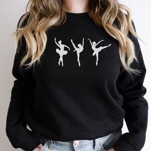 K&ouml;nnte beinhalten: Schwarzes Sweatshirt mit drei wei&szlig;en Ballettt&auml;nzerinnen-Silhouetten in verschiedenen Posen. Das Design zeigt ein klassisches Ballettthema, ideal f&uuml;r Tanzbegeisterte oder Liebhaber k&uuml;nstlerischer Kleidung.