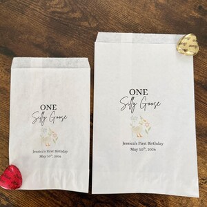 May include: Two white paper treat bags with the text "ONE Silly Goose" and "Jessica's First Birthday May 10th, 2026". The bags feature a watercolor illustration of a goose and flowers. One bag has a red heart-shaped candy, and the other has a gold-wrapped chocolate.