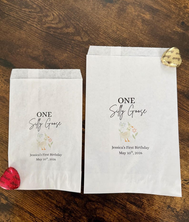 May include: Two white paper treat bags with the text "ONE Silly Goose" and "Jessica's First Birthday May 10th, 2026". The bags feature a watercolor illustration of a goose and flowers. One bag has a red heart-shaped candy, and the other has a gold-wrapped chocolate.