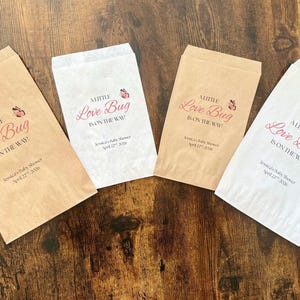 May include: Four paper favor bags, two in tan and two in white, are arranged on a dark wood surface. Each bag has the text "A Little Love Bug Is On The Way!" in pink script, with a butterfly graphic.