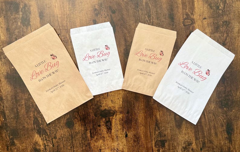 May include: Four paper favor bags, two in tan and two in white, are arranged on a dark wood surface. Each bag has the text "A Little Love Bug Is On The Way!" in pink script, with a butterfly graphic.
