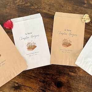 May include: Four paper favor bags, two brown and two white, with the text "A New Chapter Begins" and a floral book illustration. Each bag is sealed with a decorative heart or flower. The bags are for a baby shower.