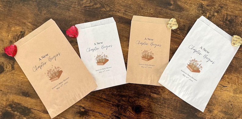 May include: Four paper favor bags, two brown and two white, with the text "A New Chapter Begins" and a floral book illustration. Each bag is sealed with a decorative heart or flower. The bags are for a baby shower.