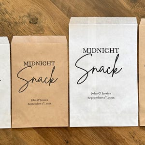 May include: Four rectangular paper bags in white and kraft brown colors, each printed with the text MIDNIGHT Snack, John & Jessica, and September 8th, 2026. A single round cookie with white frosting and dark sprinkles sits to the left of the bags.