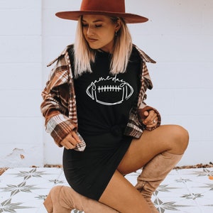 Puede incluir: Camiseta negra con la palabra "gameday" y un gr&aacute;fico de f&uacute;tbol americano en blanco. La camiseta se combina con una camisa a cuadros marr&oacute;n y un sombrero marr&oacute;n. El atuendo se completa con botas hasta la rodilla.