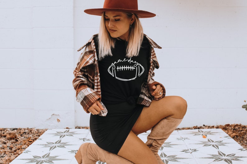 Puede incluir: Camiseta negra con la palabra "gameday" y un gr&aacute;fico de f&uacute;tbol americano en blanco. La camiseta se combina con una camisa a cuadros marr&oacute;n y un sombrero marr&oacute;n. El atuendo se completa con botas hasta la rodilla.