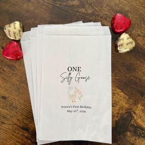 May include: White paper favor bags with the text "ONE Silly Goose" and a floral illustration. The bags are personalized with the text "Jessica's First Birthday May 10th, 2026". Gold and red foil-wrapped heart-shaped chocolates are scattered around the bags.