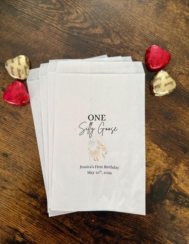 May include: White paper favor bags with the text "ONE Silly Goose" and a floral illustration. The bags are personalized with the text "Jessica's First Birthday May 10th, 2026". Gold and red foil-wrapped heart-shaped chocolates are scattered around the bags.