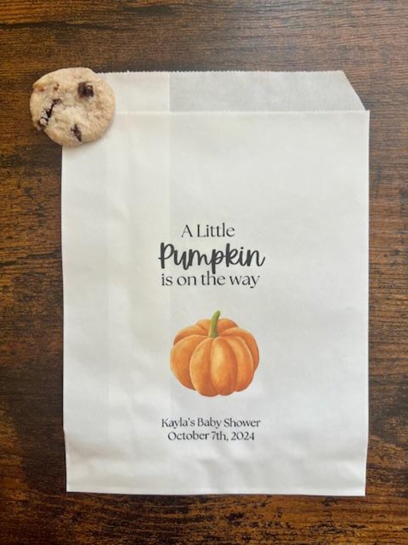 May include: White paper bag with the text "A Little Pumpkin is on the way" and a watercolor illustration of an orange pumpkin. The bag is personalized with "Kayla's Baby Shower October 7th, 2024". A chocolate chip cookie rests on the bag.