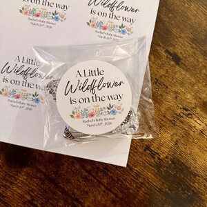May include: White circular stickers with the text "A Little Wildflower is on the way" and floral accents. The stickers are for Rachel's Baby Shower, March 20th, 2026. One sticker is on a clear plastic bag with wrapped candies.
