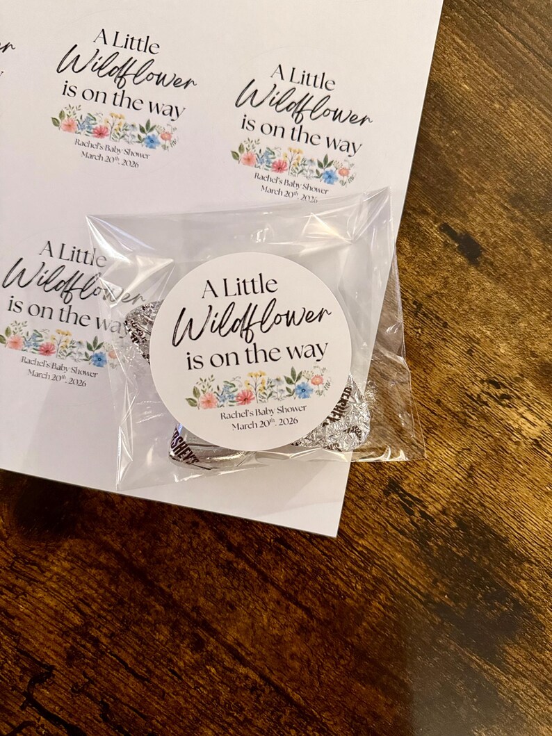 May include: White circular stickers with the text "A Little Wildflower is on the way" and floral accents. The stickers are for Rachel's Baby Shower, March 20th, 2026. One sticker is on a clear plastic bag with wrapped candies.