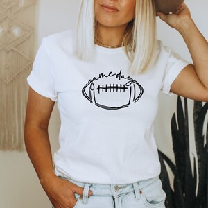 Puede incluir: Camiseta blanca con un dise&ntilde;o de f&uacute;tbol negro y la frase "game day". La camiseta tiene mangas cortas y cuello redondo. La modelo lleva vaqueros azules.
