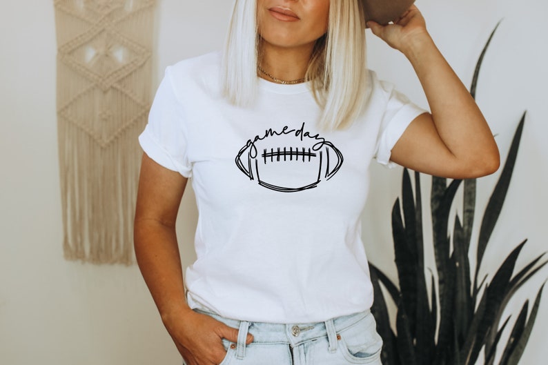Puede incluir: Camiseta blanca con un dise&ntilde;o de f&uacute;tbol negro y la frase "game day". La camiseta tiene mangas cortas y cuello redondo. La modelo lleva vaqueros azules.