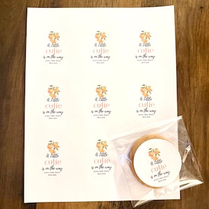 May include: A white sheet featuring eight printed circular stickers with an orange fruit illustration and the text A Little cutie is on the way, Jessica's Baby Shower, 08.02.2026. One identical sticker is applied to a round cookie in a clear bag.