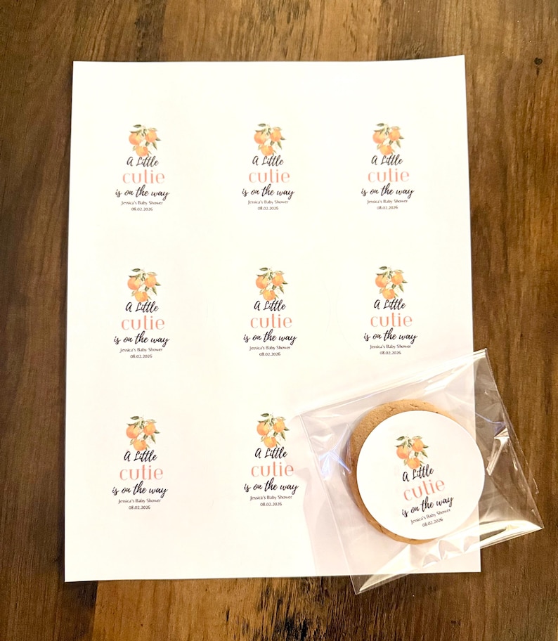 May include: A white sheet featuring eight printed circular stickers with an orange fruit illustration and the text A Little cutie is on the way, Jessica's Baby Shower, 08.02.2026. One identical sticker is applied to a round cookie in a clear bag.