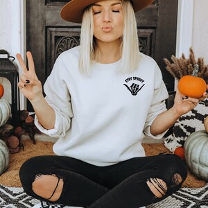 Może przedstawiać: Kremowa bluza z napisem "Stay Spooky" i grafiką szkieletu dłoni. Na zdjęciu dynie i jesienne dekoracje, sugerujące motyw Halloween. Osoba ma na sobie czarne dżinsy.