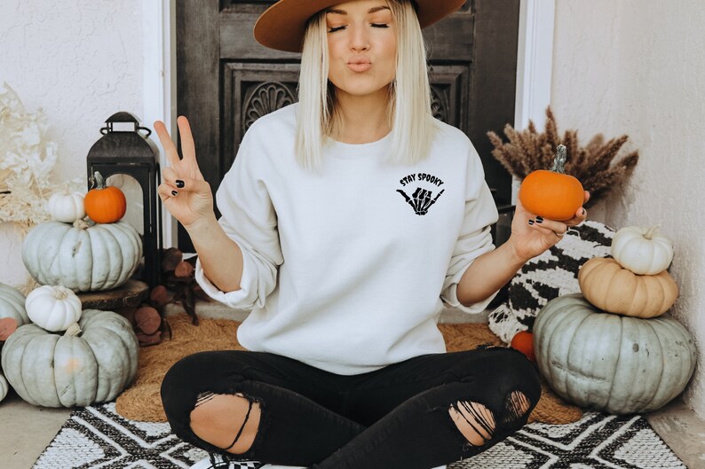 Może przedstawiać: Kremowa bluza z napisem "Stay Spooky" i grafiką szkieletu dłoni. Na zdjęciu dynie i jesienne dekoracje, sugerujące motyw Halloween. Osoba ma na sobie czarne dżinsy.