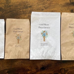 K&ouml;nnte beinhalten: Vier Papier-Geschenkt&uuml;ten in verschiedenen Gr&ouml;&szlig;en mit einem Aufdruck. Das Design zeigt den Text "God Bless Theo James" &uuml;ber einem Kreuz mit blauen Blumen. Der Text "Theo's Baptism May 26th, 2026" ist darunter gedruckt.