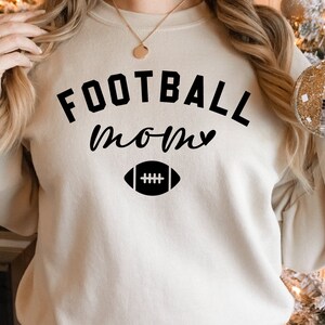 May include: A beige sweatshirt with the words "FOOTBALL MOM" in black script and a football graphic. The sweatshirt is a crew neck style and appears to be made of a soft material. The overall theme is sports and family.