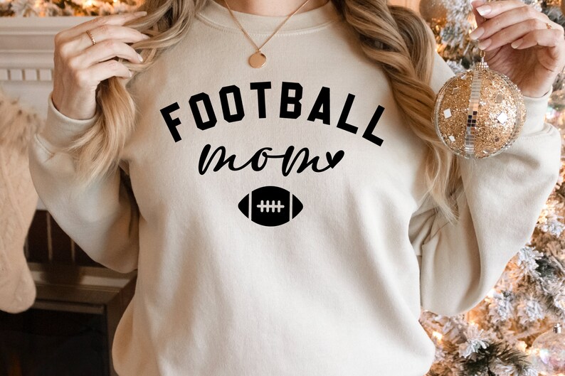 May include: A beige sweatshirt with the words "FOOTBALL MOM" in black script and a football graphic. The sweatshirt is a crew neck style and appears to be made of a soft material. The overall theme is sports and family.