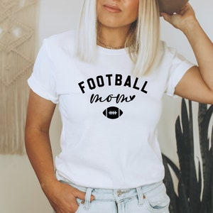 May include: White t-shirt with the words "FOOTBALL mom" in black script and a football graphic. The shirt is a classic crew neck style and is worn by a person. The shirt is paired with blue jeans.