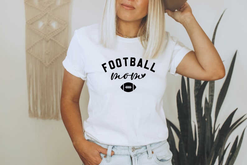May include: White t-shirt with the words "FOOTBALL mom" in black script and a football graphic. The shirt is a classic crew neck style and is worn by a person. The shirt is paired with blue jeans.