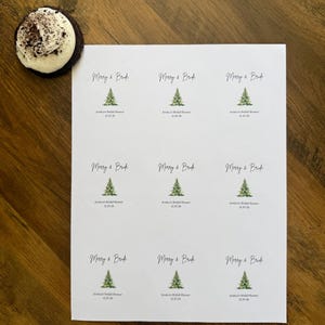 May include: A white sheet containing nine circular stickers, each featuring a green watercolor Christmas tree illustration, the text Merry & Bride, and the custom text Jessica's Bridal Shower 12.20.26. A single chocolate cookie with white frosting is nearby.