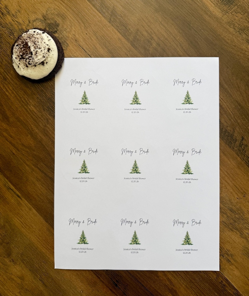May include: A white sheet containing nine circular stickers, each featuring a green watercolor Christmas tree illustration, the text Merry & Bride, and the custom text Jessica's Bridal Shower 12.20.26. A single chocolate cookie with white frosting is nearby.