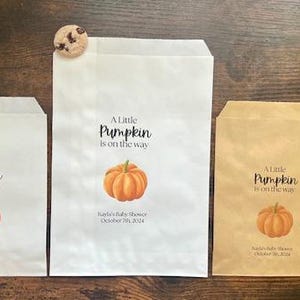 May include: Three paper treat bags with the text "A Little Pumpkin is on the way" and a pumpkin graphic. The bags are white and brown, with a cookie on each. The text "Kayla's Baby Shower October 7th, 2024" is also printed on the bags.