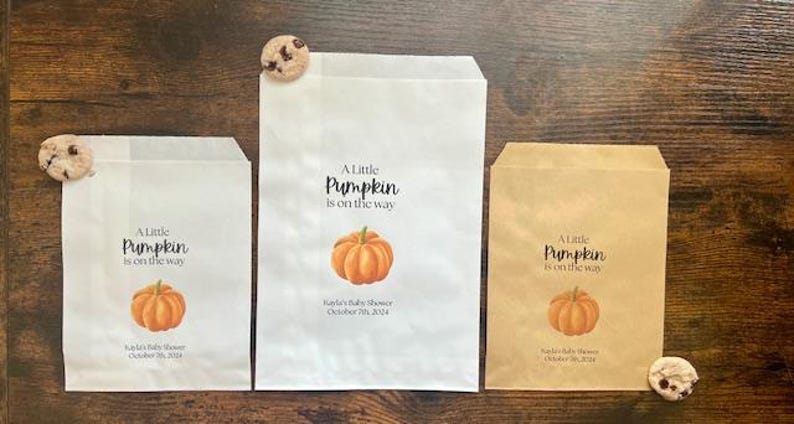May include: Three paper treat bags with the text "A Little Pumpkin is on the way" and a pumpkin graphic. The bags are white and brown, with a cookie on each. The text "Kayla's Baby Shower October 7th, 2024" is also printed on the bags.