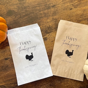 May include: Thanksgiving-themed gift bags, one white and one brown, with a black turkey silhouette and the words "Happy Thanksgiving!" printed on them. An orange pumpkin and a white pumpkin are also in the image.