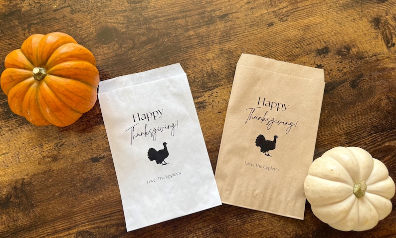 May include: Thanksgiving-themed gift bags, one white and one brown, with a black turkey silhouette and the words "Happy Thanksgiving!" printed on them. An orange pumpkin and a white pumpkin are also in the image.