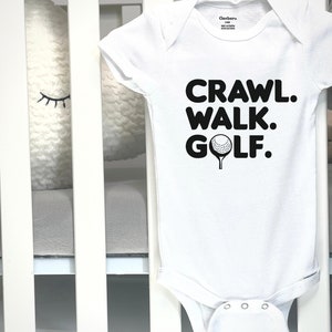May include: White baby bodysuit featuring the words "CRAWL. WALK. GOLF." in black, and a golf ball graphic. The garment is shown in a cot with white bars and a grey mattress.
