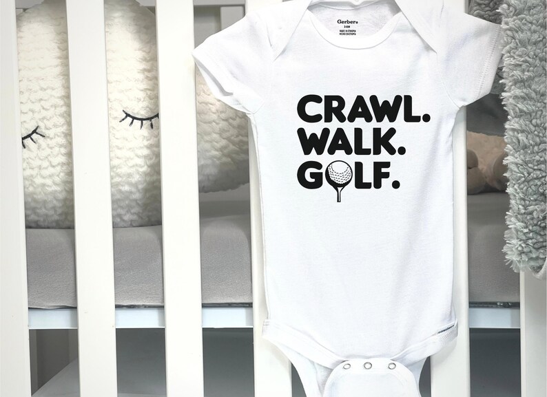 May include: White baby bodysuit featuring the words "CRAWL. WALK. GOLF." in black, and a golf ball graphic. The garment is shown in a cot with white bars and a grey mattress.