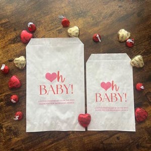 May include: Two white paper gift bags with the text "Oh Baby!" in pink, surrounded by heart-shaped chocolates. The bags also include the text "A little sweetheart is on the way. Thank you for showering Jessica."