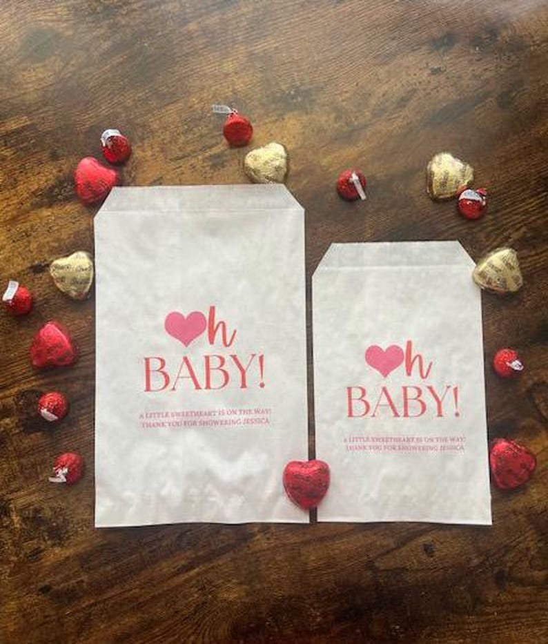 May include: Two white paper gift bags with the text "Oh Baby!" in pink, surrounded by heart-shaped chocolates. The bags also include the text "A little sweetheart is on the way. Thank you for showering Jessica."